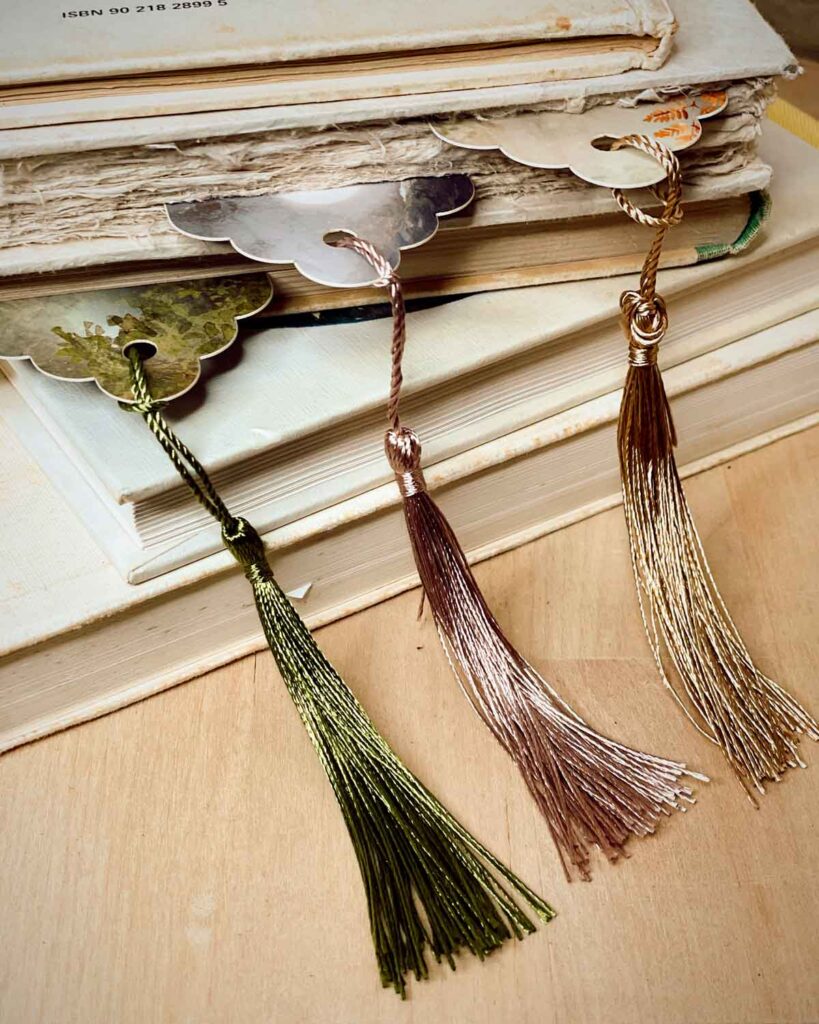 Three scalloped bookmarks with shimmering tassels in forest green, rose gold, and bronze, peeking out from vintage books.