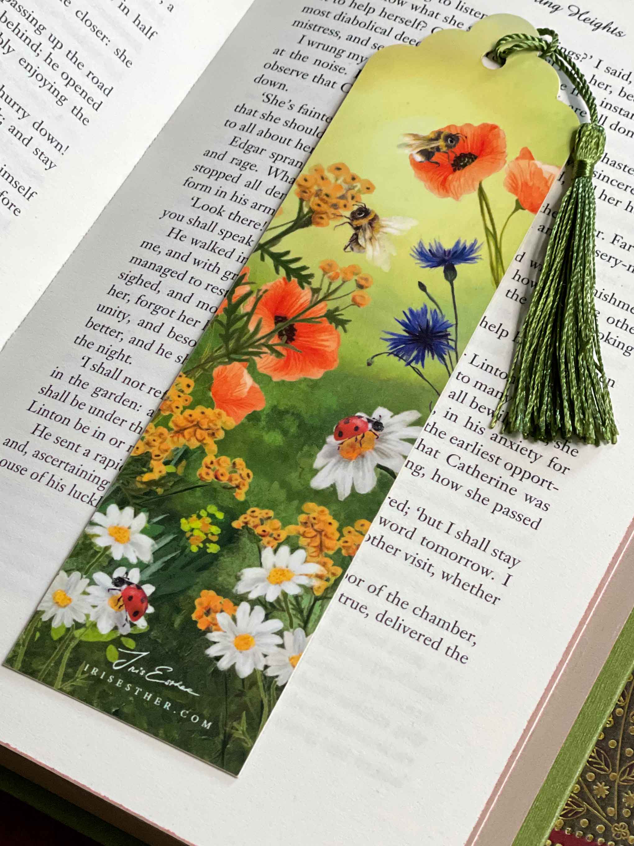 Illustrated bookmark featuring a vibrant wildflower meadow with poppies, daisies, cornflowers, and bees, finished with a green tassel, resting on an open book.