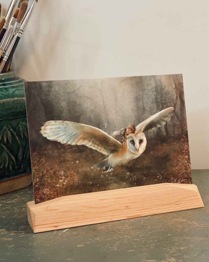 Watercolor painting of barn owl in flight through misty forest displayed in wooden stand on artist's desk